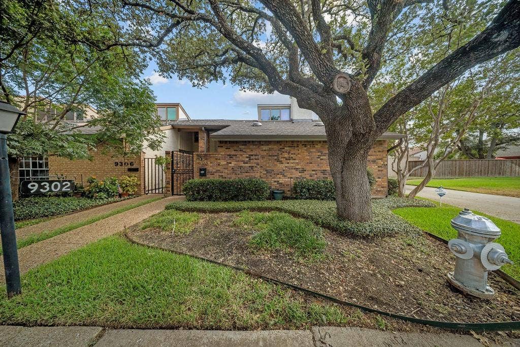 9302 Chimney Sweep Lane, Dallas, TX 75243 Brick facade residence with a mature front yard tree, a gravel pathway, and an ornamental security gate