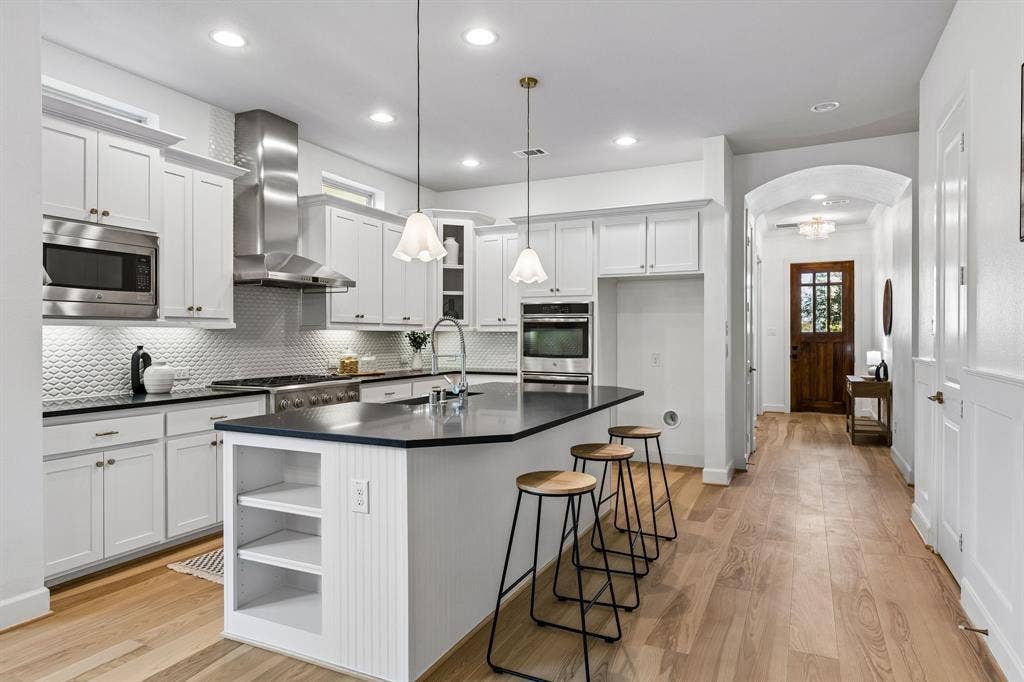2221 Tremont Boulevard, McKinney, TX 75071 Kitchen featuring white shaker-style cabinetry, stainless steel appliances, dark countertops, a large island with shelving, and wood-finish flooring