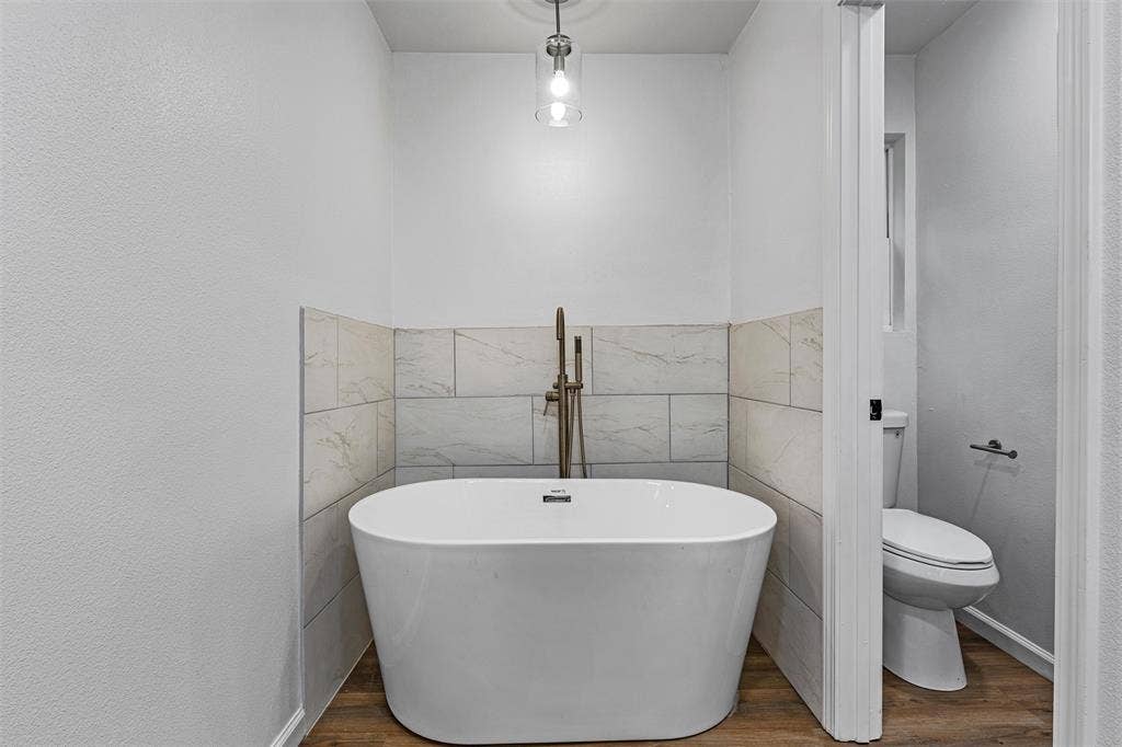 1132 Western Oaks Drive, Woodway, TX 76712 Freestanding soaking tub with floor-mounted faucet, tile wainscoting, wood-finish flooring, and a contemporary pendant light fixture