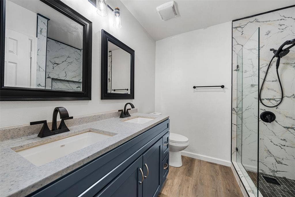 1132 Western Oaks Drive, Woodway, TX 76712 Dual vanity with integrated sinks and dark blue cabinetry, quartz-finish countertop, black-framed mirrors, and a glass-enclosed shower with a marble-patterned tile surround