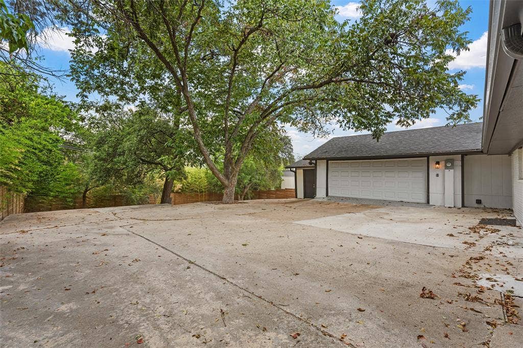1132 Western Oaks Drive, Woodway, TX 76712 Expansive concrete driveway leading to a multi-car garage with a panel door, accompanied by mature trees and a privacy fence