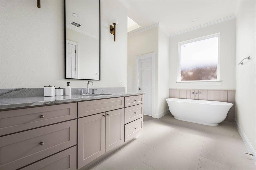 5901 Glen Heather Drive, Plano, TX 75093 Serene primary bath featuring a freestanding soaking tub beneath a large window, separate vanity, large-format tile flooring, tailored cabinetry, and understated contemporary finishes.
