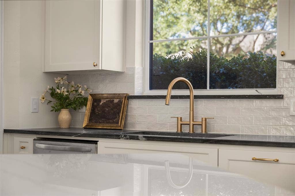 5901 Glen Heather Drive, Plano, TX 75093 Thoughtfully selected finishes include dark perimeter countertops, a brass-toned bridge faucet, textured white backsplash tile, stainless-steel appliances, and cabinetry extending around the window for additional storage.