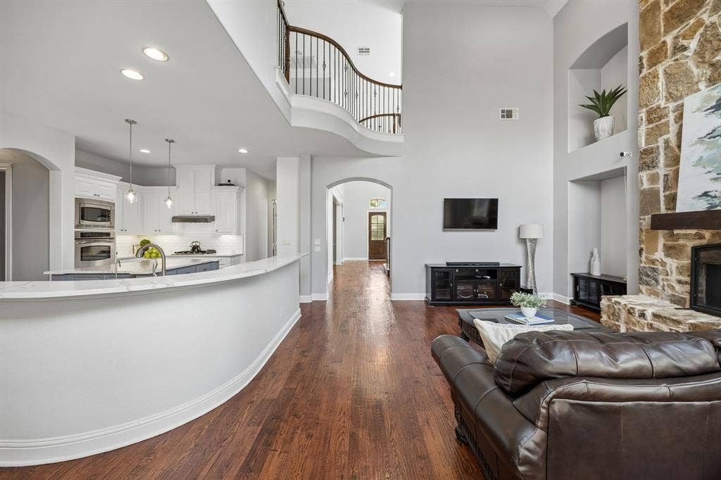 1011 Three Rivers Drive, Prosper, TX 75078 Light-filled living area with soaring ceilings, rich wood floors, and seamless flow to the kitchen, anchored by a striking stone fireplace.