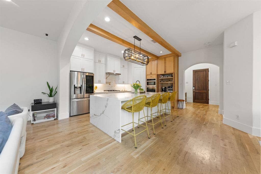 7011 Nueces Drive, Irving, TX 75039 Gourmet kitchen featuring a large waterfall island with seating, stainless steel appliances, wood-finish flooring, white cabinetry, and wood beam accents