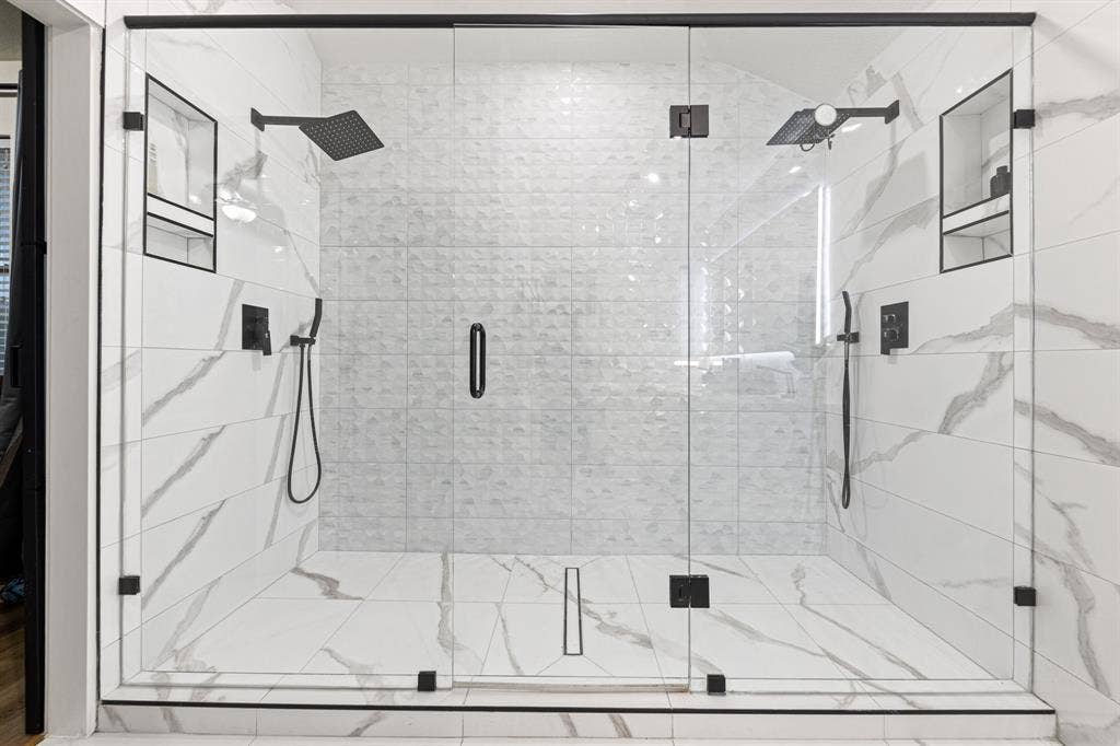 7011 Nueces Drive, Irving, TX 75039 Glass-enclosed shower with contrasting white and textured wall tiles, dual rainfall showerheads, and integrated wall niches