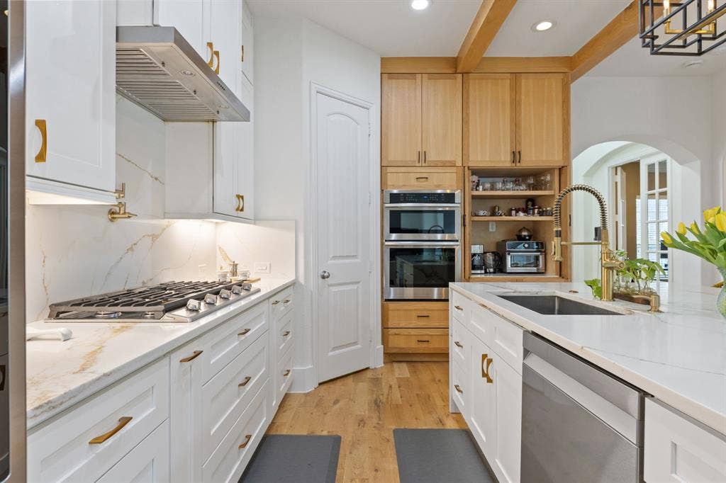 7011 Nueces Drive, Irving, TX 75039 White shaker cabinetry with gold-tone hardware, stainless steel gas cooktop, double wall ovens, wood-finish flooring, and a kitchen island with an undermount sink