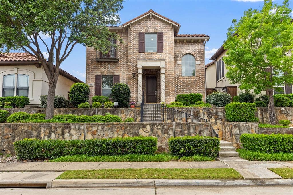 7011 Nueces Drive, Irving, TX 75039 Brick facade with stone accents, arched windows, covered entry, and tiered landscaping with mature trees and shrubs