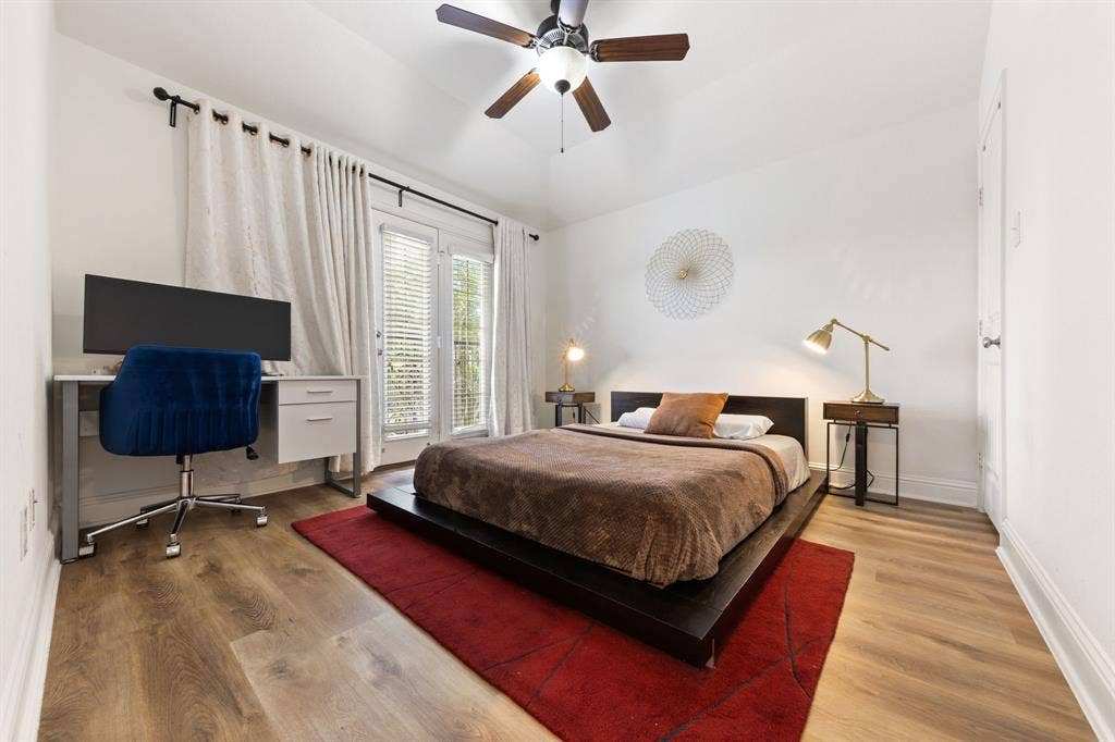 7011 Nueces Drive, Irving, TX 75039 Spacious bedroom featuring hardwood flooring, a ceiling fan with integrated lighting, and large windows with plantation shutters and layered curtains providing natural light and privacy
