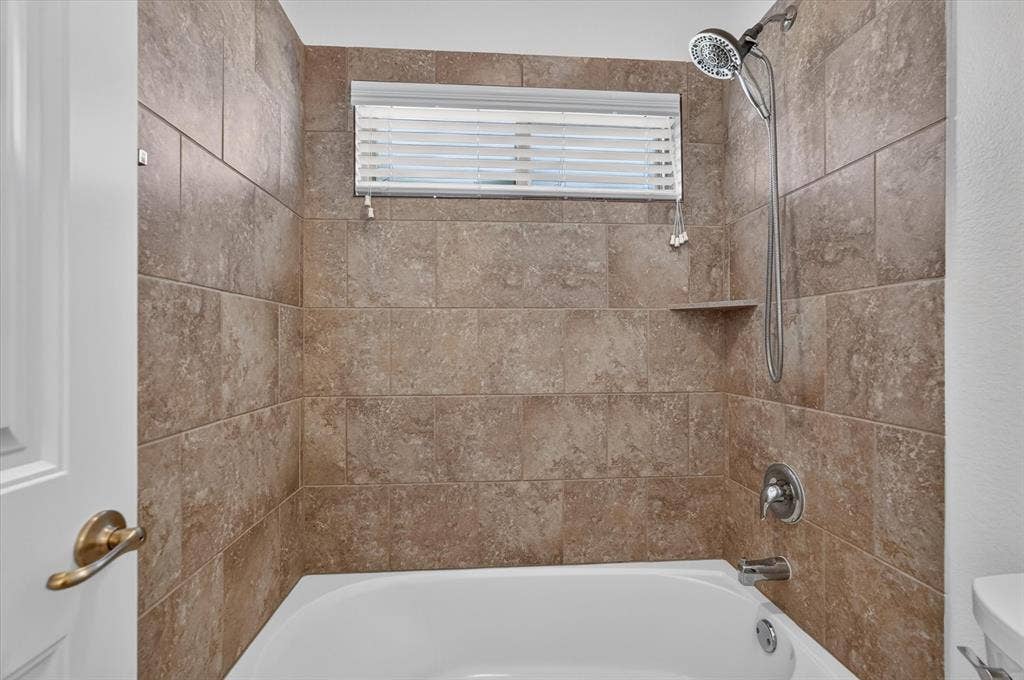 Bathtub with full-height tiled surround, integrated shower niche, and a window with blinds