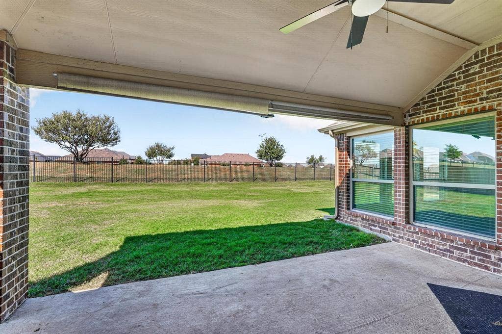 Covered concrete patio with a ceiling fan, brick pillars, and expansive lawn beyond a metal fence