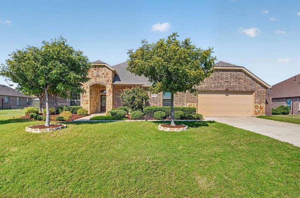Brick and stone facade, arched entryway, attached garage, concrete driveway, and landscaped front yard with mature trees