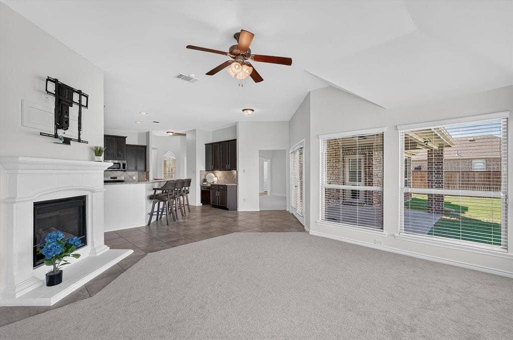 Vaulted ceiling with ceiling fan, white fireplace with hearth, kitchen with breakfast bar, built-in cabinetry, and multiple windows with blinds