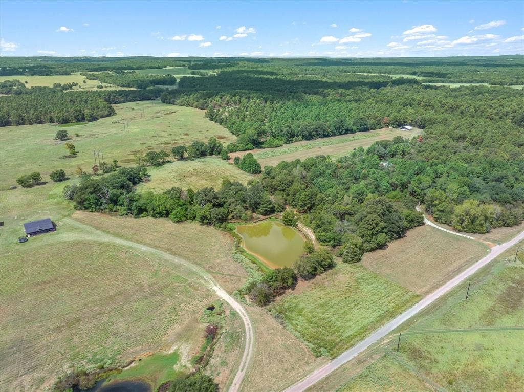 24619 Private Road 8102, Frankston, TX 75763 Expansive rural landscape featuring a pond, wooded areas, open fields, a paved road, and a black-roofed structure