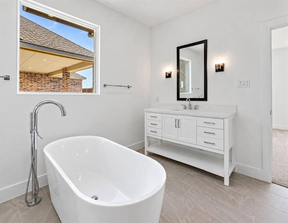 1411 Potenza Road, Fate, TX 75087 Freestanding soaking tub with a floor-mounted chrome faucet, white vanity with dual cabinet doors and multiple drawers, framed mirror, and wall-mounted sconces