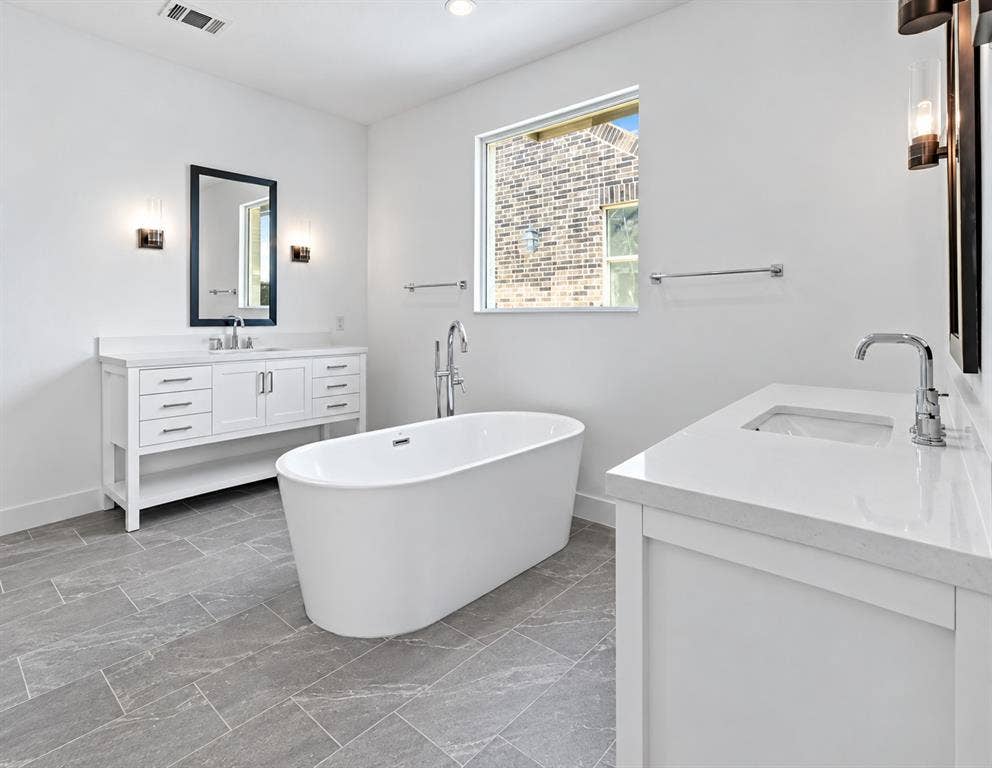 1411 Potenza Road, Fate, TX 75087 Freestanding soaking tub, dual white vanity cabinets, white countertops, chrome fixtures, and gray rectangular floor tiling