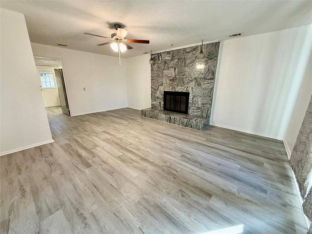 Spacious room featuring wood-finish flooring, a stone fireplace with hearth, and a ceiling fan with light fixture
