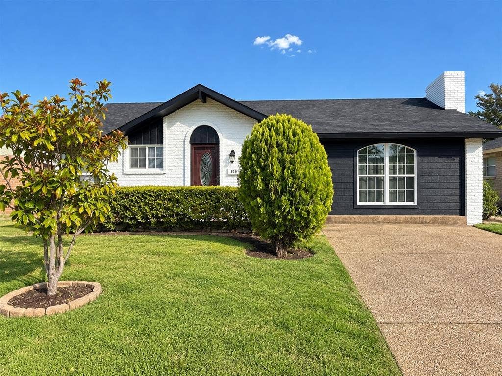 White brick exterior with dark shingle roofing, an arched entryway, a prominent arched window, and a light-colored driveway
