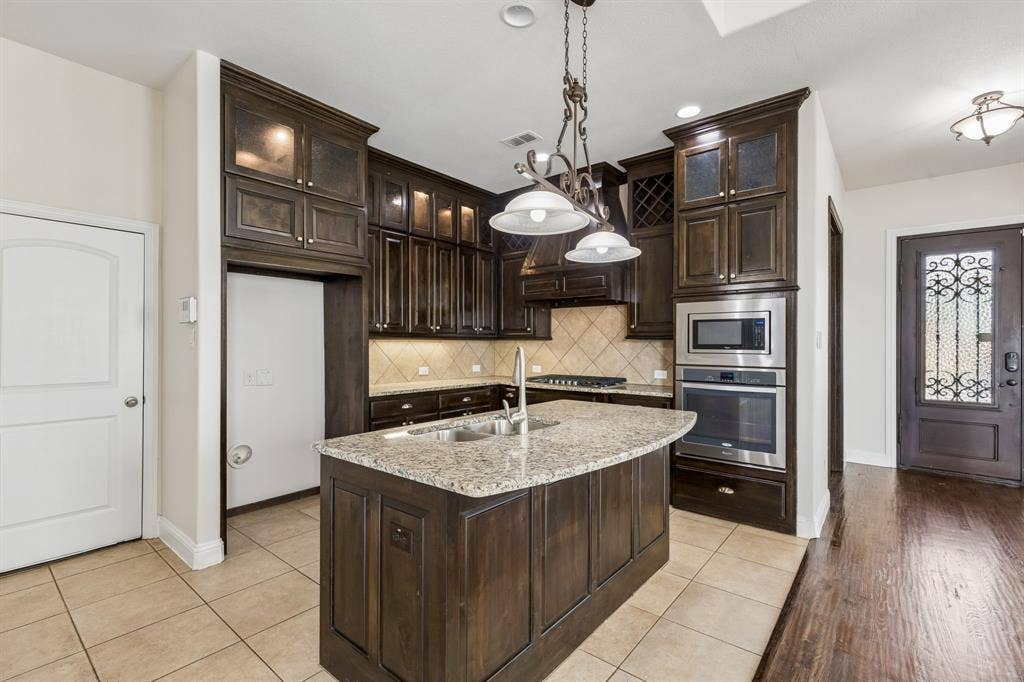 Kitchen featuring dark wood cabinetry, a central island with a granite countertop and sink, stainless steel appliances including a built-in oven and microwave, and a tiled backsplash