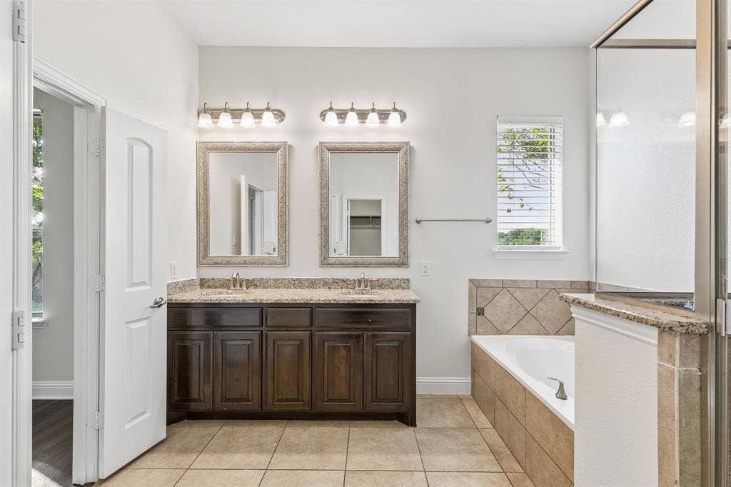 Dual vanity with dark wood-finish cabinetry and stone-finish countertops, framed mirrors, tiled flooring, and a tiled tub surround
