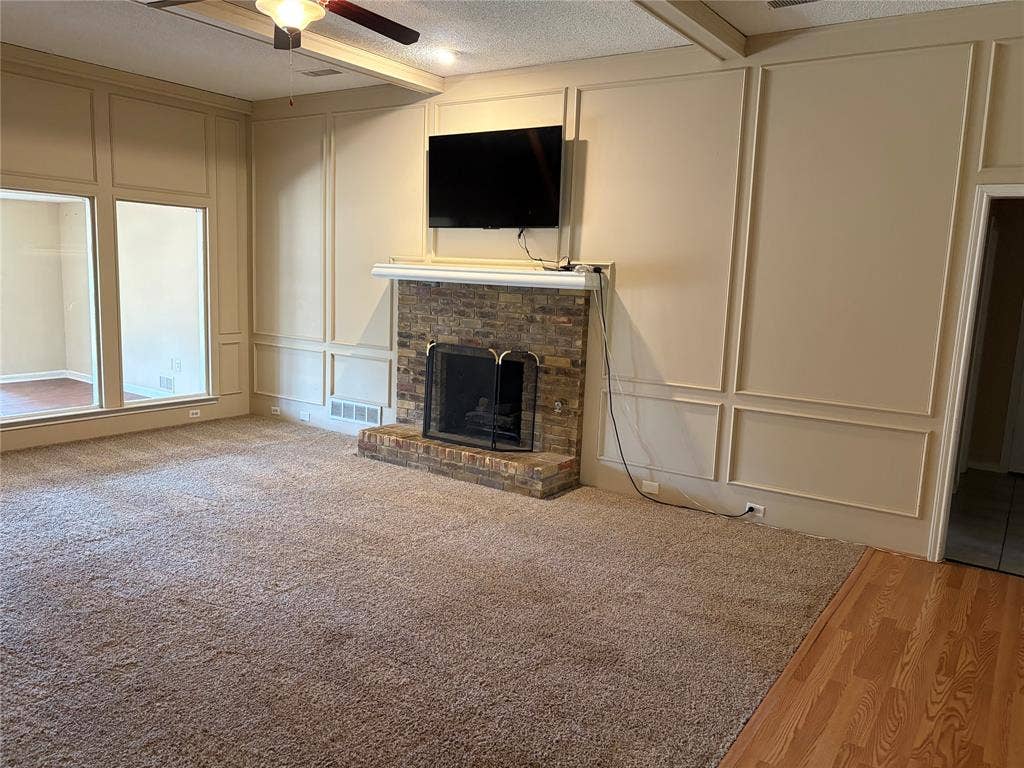 3912 Bandera Drive, Plano, TX 75074 Stone-faced fireplace with a white mantel, large picture windows, decorative wall paneling, ceiling fan with light fixture, and wood-finish flooring