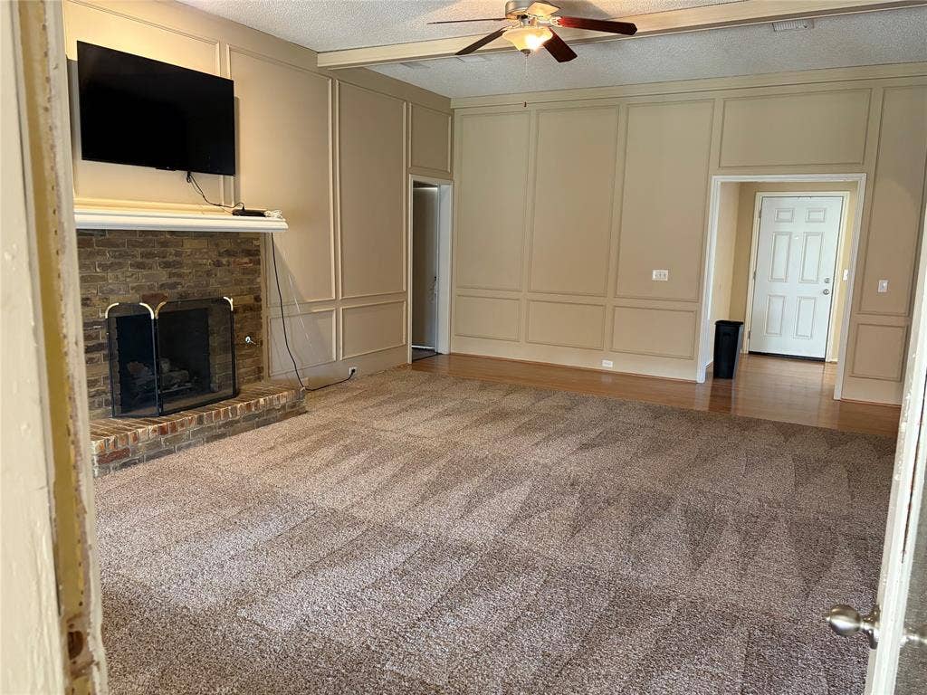 3912 Bandera Drive, Plano, TX 75074 Living area featuring a brick fireplace with a white mantel, decorative wall paneling, a ceiling fan with integrated lighting, and a mix of carpeted and wood-finish flooring