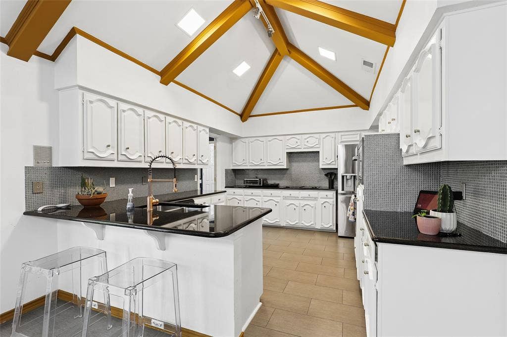 5716 Plumtree Drive, Dallas, TX 75252 Vaulted ceiling with exposed wood beams, white cabinetry, dark countertops, gray mosaic tile backsplash, and light wood-finish flooring