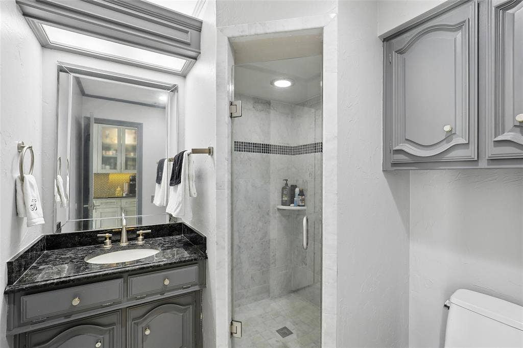 5716 Plumtree Drive, Dallas, TX 75252 Adjacent to family room. Vanity with dark granite countertop and undermount sink, framed mirror with overhead lighting, glass-enclosed shower with tile surround, and built-in cabinetry