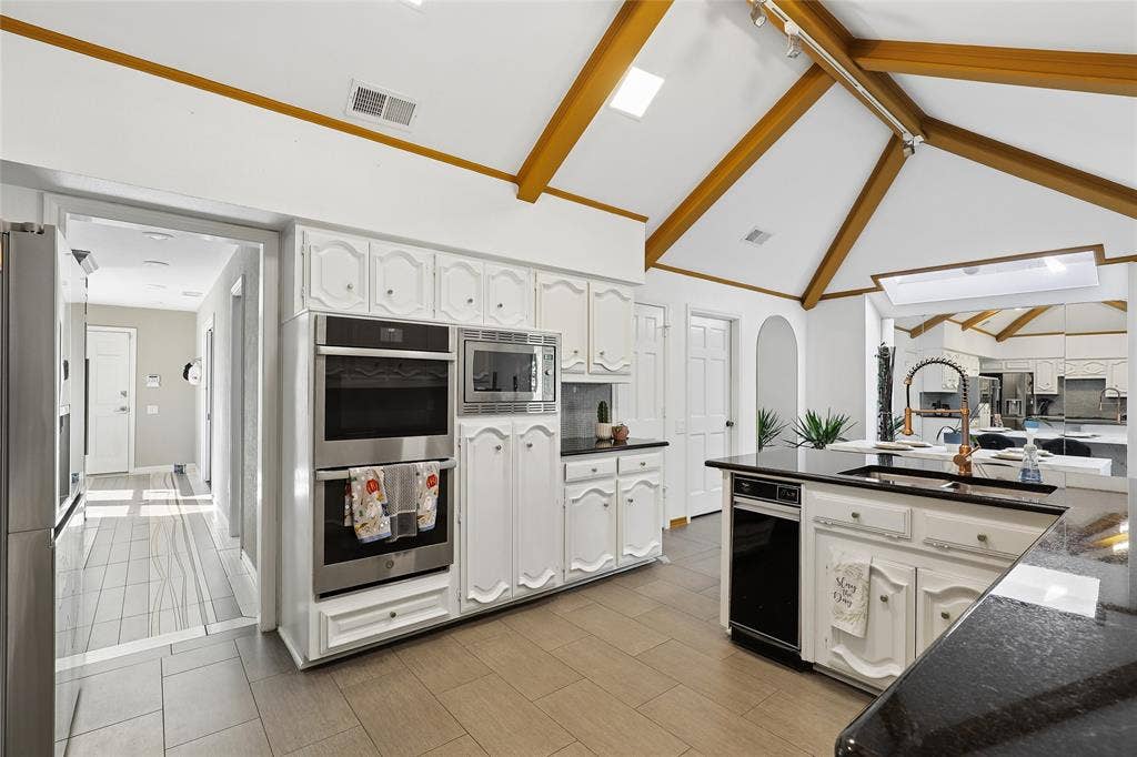 5716 Plumtree Drive, Dallas, TX 75252 Vaulted ceiling with exposed wood beams, stainless steel double wall ovens, built-in microwave, white cabinetry, and dark granite countertops