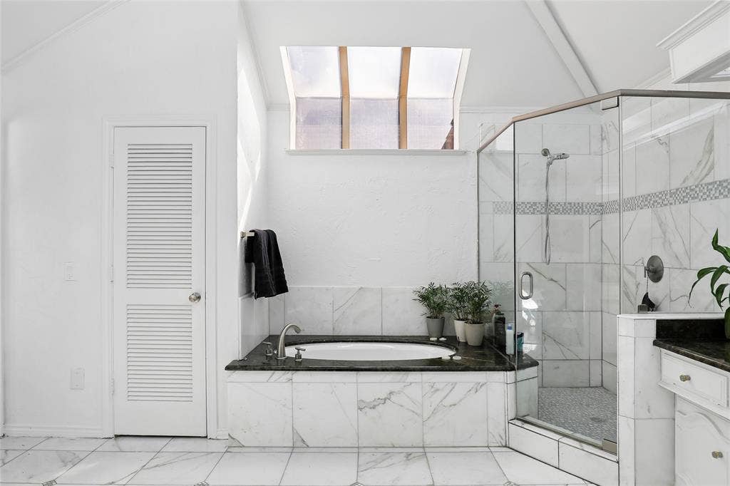 5716 Plumtree Drive, Dallas, TX 75252 Recessed bathtub with dark stone surround, walk-in shower with clear glass enclosure, white marble-style wall and floor tiling, and a ceiling skylight