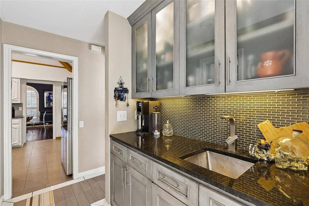 5716 Plumtree Drive, Dallas, TX 75252 Wet bar with gray cabinetry, luxury granite, frosted glass upper cabinet doors, and a hammered copper sink. Also includes huge wine fridge and custom tile flooring.
