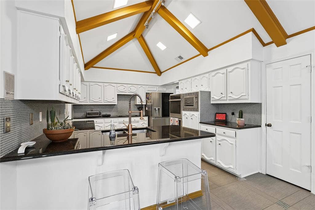 5716 Plumtree Drive, Dallas, TX 75252 Vaulted ceiling with exposed beams, white cabinetry, dark granite countertops, mosaic tile backsplash, and built-in stainless-steel appliances