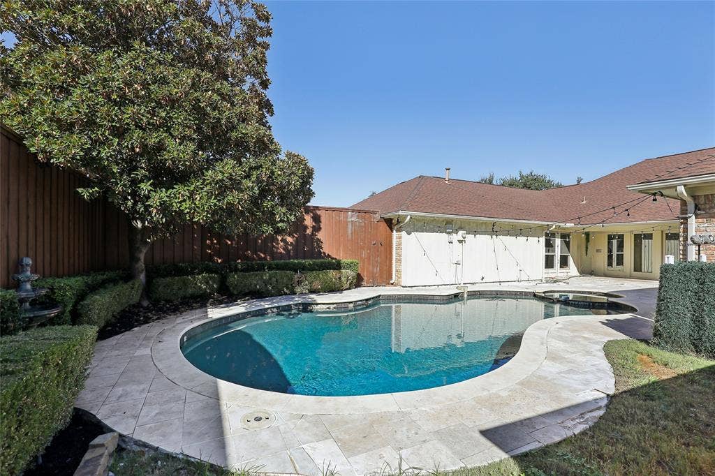 5716 Plumtree Drive, Dallas, TX 75252 Curved swimming pool with an integrated spa, surrounded by stone-finish coping and a paved patio