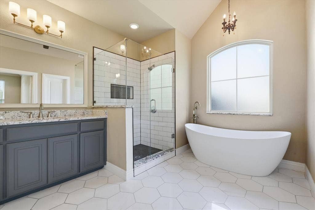 3110 Creek Haven Drive, Highland Village, TX 75077 Freestanding soaking tub beneath an arched window, dual-sink vanity with stone countertop, and walk-in shower featuring a glass enclosure and subway tile surround, finished with hexagonal floor tile and vaulted ceilings