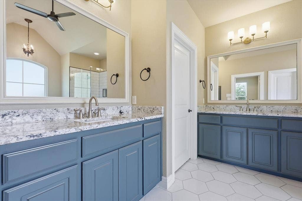 3110 Creek Haven Drive, Highland Village, TX 75077 Dual vanity featuring blue cabinetry, patterned stone-finish countertops, rectangular undermount sinks, large framed mirrors, and white hexagonal floor tiling