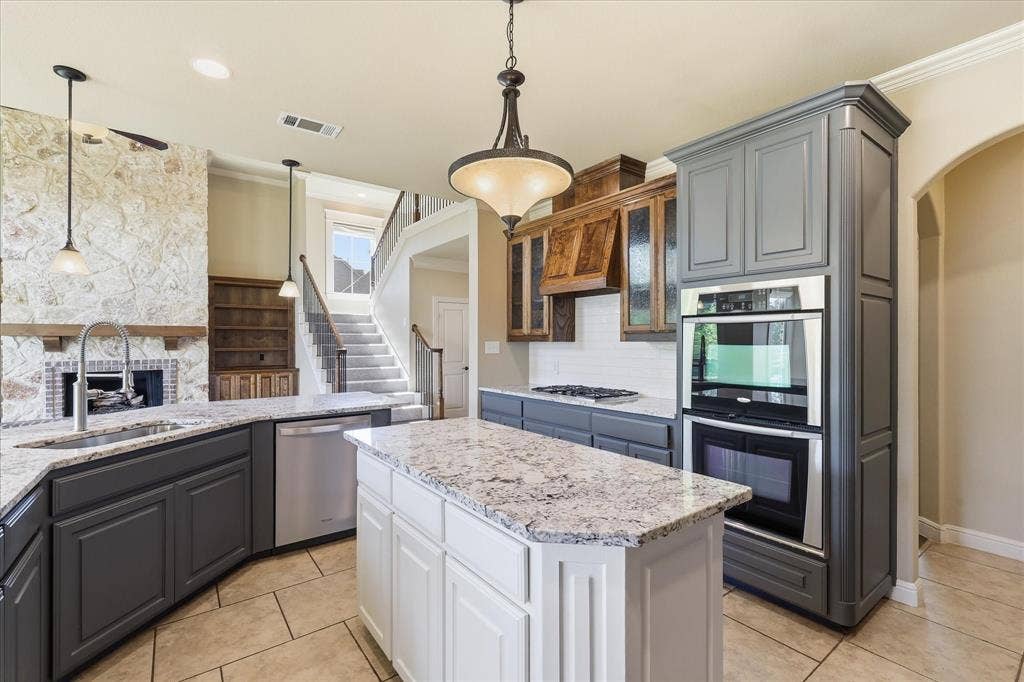3110 Creek Haven Drive, Highland Village, TX 75077 Kitchen featuring a stone-faced fireplace, center island with stone countertop, stainless steel double wall ovens, two-tone cabinetry, and wood-finish cabinetry accents