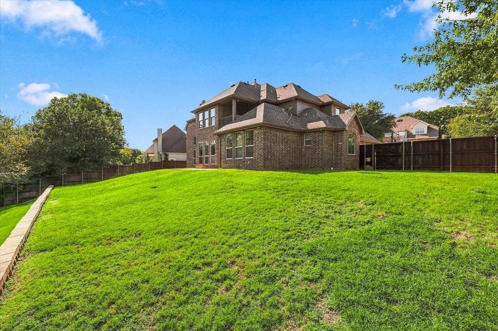 3110 Creek Haven Drive, Highland Village, TX 75077 Brick exterior two-story residence featuring an expansive grass lawn, tiered stone retaining wall, and wood privacy fencing