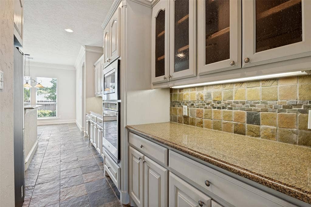 806 Cobblestone Court, Cedar Hill, TX 75104 White cabinetry with glass-front upper cabinets, light-toned stone tile flooring, built-in stainless steel oven, stone tile backsplash, and granite-finish countertops