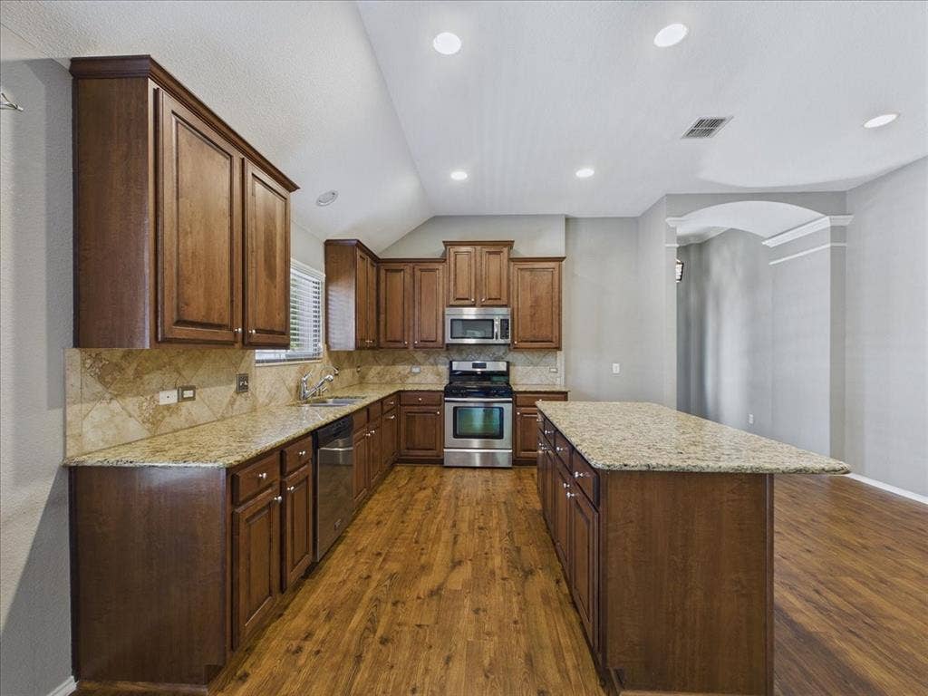 Kitchen featuring wood-finish cabinetry, granite countertops, tile backsplash, stainless steel appliances, and wood-finish flooring