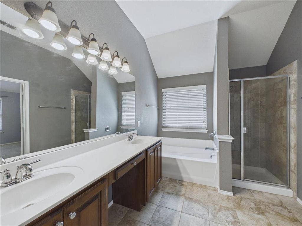 Dual vanity with integrated sinks, wood-finish cabinetry, built-in bathtub, glass-enclosed shower, and tiled flooring