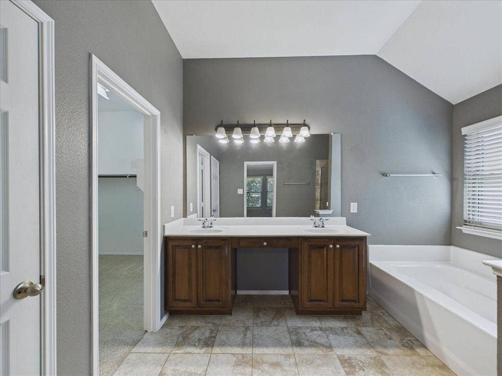 Spa-inspired primary bathroom featuring a dual-sink vanity with wood cabinetry, a large framed mirror with integrated lighting, a soaking tub, a walk-in shower with glass enclosure, tile flooring, and abundant natural light from multiple windows