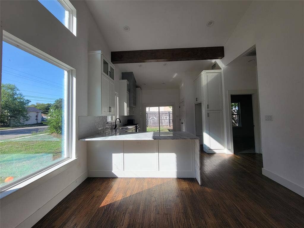 Open-concept kitchen featuring wood-finish flooring, white cabinetry, stone-finish countertops, exposed ceiling beam, and oversized windows