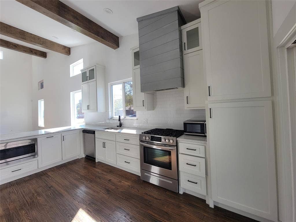 Kitchen featuring exposed timber ceiling beams, shaker-style cabinetry, a custom range hood, and stainless-steel appliances atop wood-finish flooring
