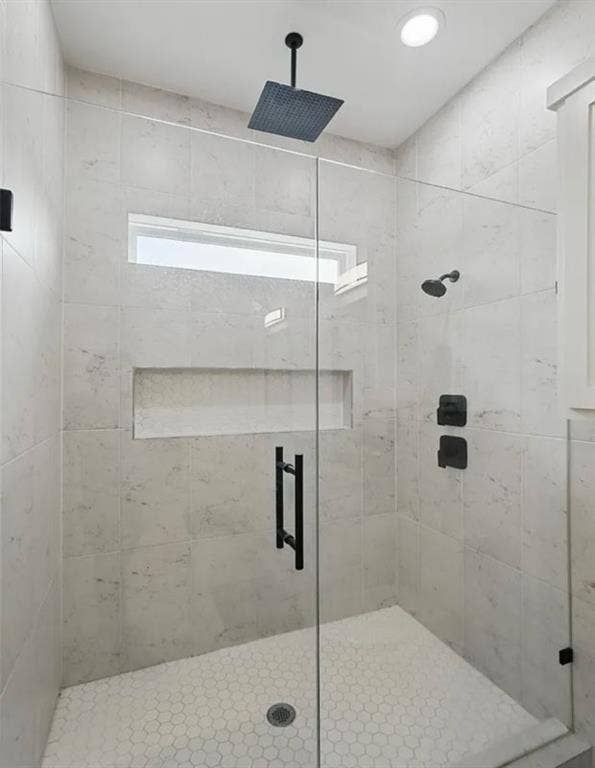Walk-in shower featuring large-format stone-look wall tiles, white hexagonal floor tiling, a frameless glass enclosure, built-in recessed shelving, and matte black rainfall and handheld shower fixtures