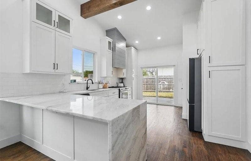Kitchen featuring white shaker-style cabinetry, stone-surface countertops with waterfall edge, wood-finish flooring, a custom range hood, and an exposed wooden ceiling beam