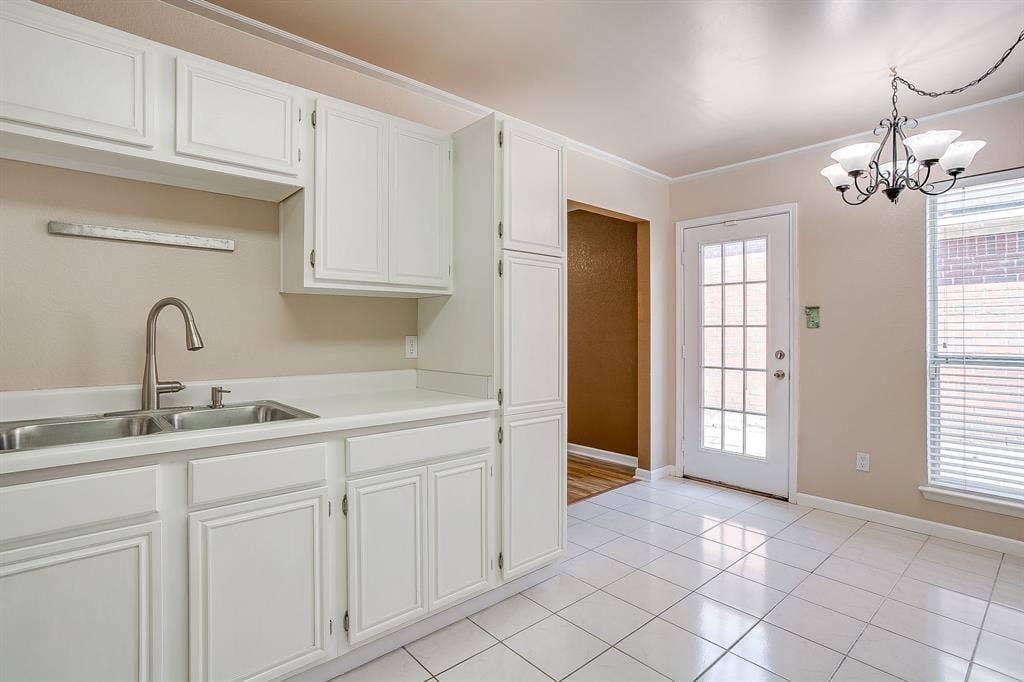 White shaker-style cabinetry, double-basin stainless steel sink with high-arc faucet, white tile flooring, glass-paneled exterior door, and decorative multi-arm chandelier
