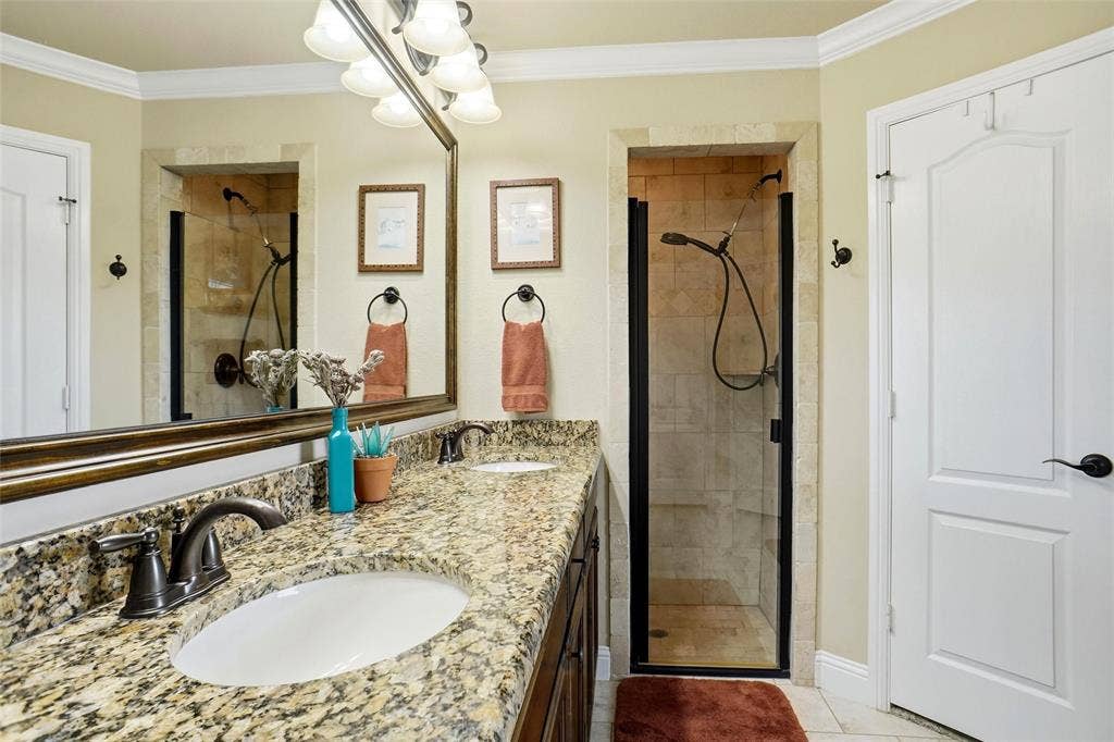 Dual-sink vanity with stone countertops and bronze-finish fixtures, framed wall mirror, and walk-in shower featuring tile surround and a dark-finish door frame