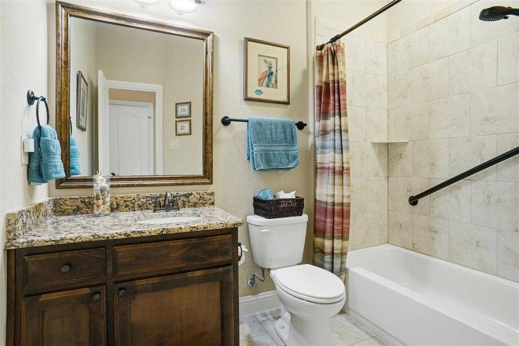 Bathroom featuring a vanity with stone countertop, wood cabinetry, framed mirror, and tiled shower surround with a metal grab bar