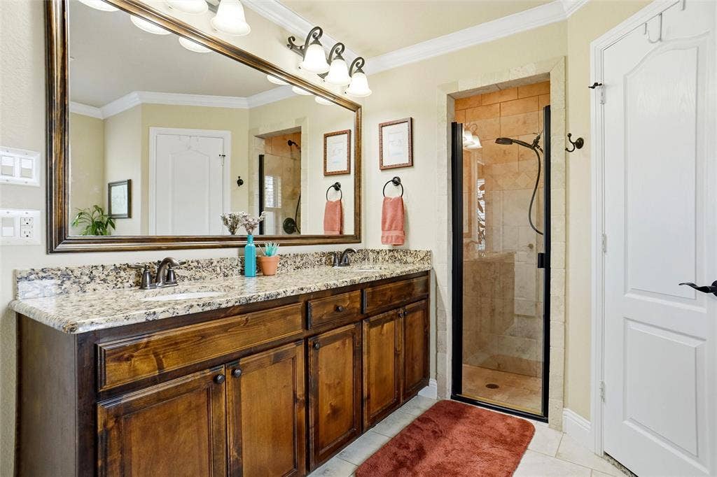 Dual-sink vanity with stone-finish countertop, dark wood-finish cabinetry, oversized framed wall mirror, and walk-in shower featuring glass enclosure and stone-finish wall tile