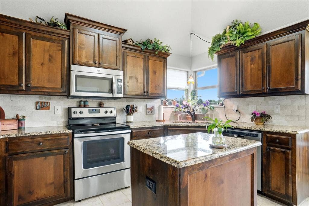 Kitchen featuring stained wood cabinetry, stone countertops, stone tile backsplash, stainless steel appliances, and a central island